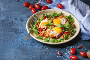 Traditional Israeli Cuisine dishes Shakshuka. Fried egg with vegetables tomatoes and paprika in ceramic plate with cloth, herbs and ingredients above over blue texture background. Copy space.