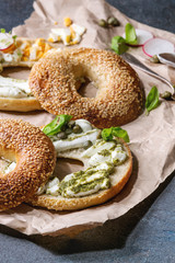 Variety of homemade bagels with sesame seeds, cream cheese, pesto sauce, eggs, radish, herbs served on crumpled paper with ingredients above over blue texture background. Close up
