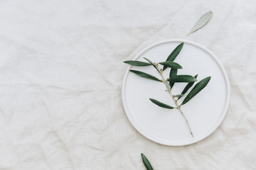 Flat lay olive branch on white plate over linen tablecloth. Organic rustic background with copy...