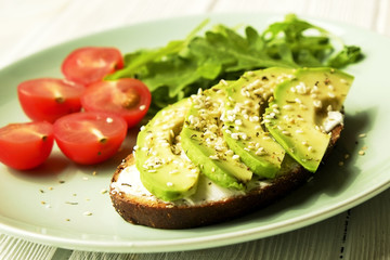 Vegan sandwich, rye bread toast, avocado, vegenaise eggless mayonnaise sauce, sesame seeds, cherry tomato arugula rucola leaves, plate, wooden table. National avocado day concept. Close up, background