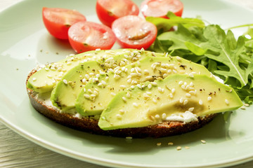 Vegan sandwich, rye bread toast, avocado, vegenaise eggless mayonnaise sauce, sesame seeds, cherry tomato arugula rucola leaves, plate, wooden table. National avocado day concept. Close up, background