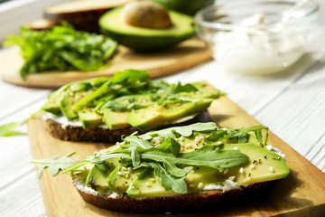 Vegan sandwich, rye bread toast, avocado, vegenaise eggless mayonnaise sauce, sesame seeds seasoning, arugula rucola leaves white wood texture table. National avocado day concept. Close up, background