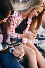 Two beautiful girls in colored shirts sitting on the grass on the hillside. Watch the sunset, drinking wine, hug, happy