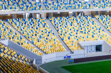 Fototapeta premium Siedzący kibice kolorowe plastikowe krzesła na tle stadionu piłkarskiego. Pusty stadion boisko do piłki nożnej zielona trawa na arenie lekkoatletycznej piłki nożnej.