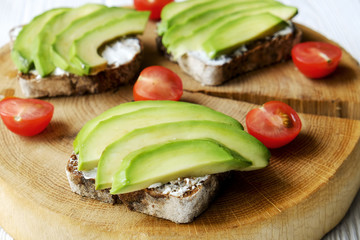 Vegan sandwich, rye bread toast, avocado, cherry tomato, vegenaise eggless mayonnaise sauce on thick cracked wood slice, white wooden texture table. National avocado day concept. Close up, background.