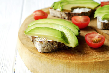 Vegan sandwich, rye bread toast, avocado, cherry tomato, vegenaise eggless mayonnaise sauce on thick cracked wood slice, white wooden texture table. National avocado day concept. Close up, background.