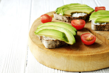 Vegan sandwich, rye bread toast, avocado, cherry tomato, vegenaise eggless mayonnaise sauce on thick cracked wood slice, white wooden texture table. National avocado day concept. Close up, background.
