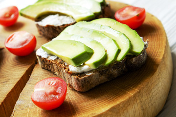 Vegan sandwich, rye bread toast, avocado, cherry tomato, vegenaise eggless mayonnaise sauce on thick cracked wood slice, white wooden texture table. National avocado day concept. Close up, background.