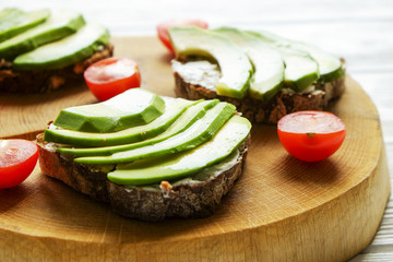Vegan sandwich, rye bread toast, avocado, cherry tomato, vegenaise eggless mayonnaise sauce on thick cracked wood slice, white wooden texture table. National avocado day concept. Close up, background.