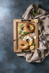 Variety of homemade bagels with sesame seeds, cream cheese, pesto sauce, eggs, radish, herbs served on wooden tray with cloth and ingredients above over blue texture background. Top view, space.