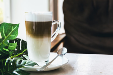 A cup of Hot cappuccino latte coffee with cinnamon sprinkles on top on wooden white dinner table with a green leaf in a  background