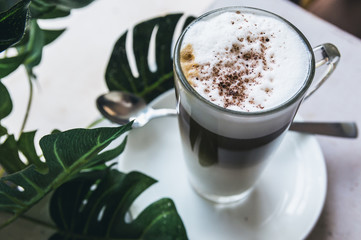 A cup of Hot cappuccino latte coffee with cinnamon sprinkles on top on wooden white dinner table with a green leaf in a  background