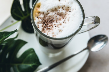 A cup of Hot cappuccino latte coffee with cinnamon sprinkles on top on wooden white dinner table with a green leaf in a  background