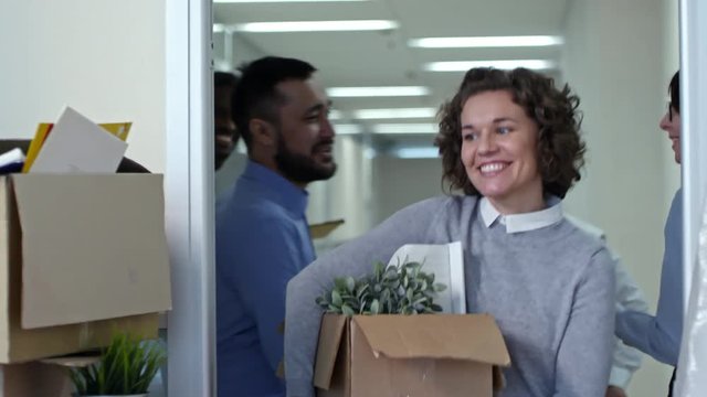 Joyous woman carrying cardboard box and taking key while men bringing furniture into new office