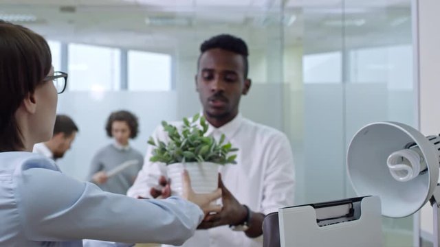 Young businesswoman unpacking belongings with assistance of male african american colleague after moving in new office; group of coworkers taking off stuff from box in background