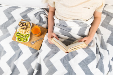 Reading book in bed and having breakfast, top view. Flat lay of female person in clean white minimalistic bed at home or hospital with healthy vegan diet breakfast and juice glass