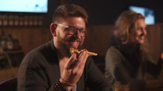 Young man with beard and glasses is bringing piece of pizza from plate to face. He is resting in a pizzeria with company of friends