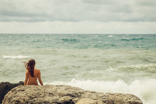 Naked young girl with a beautiful body stands facing the sea, back to the beach and watching the raging, mighty, strong waves during a storm in the ocean. Beach and stones. 