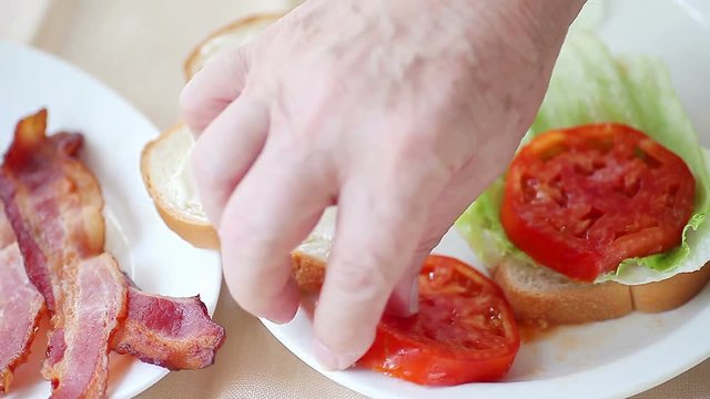 A man adds tomato and bacon slices to lettuce on white bread with mayonnaise