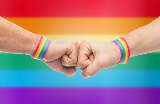 lgbt, same-sex love and homosexual relationships concept - close up of male couple hands with gay pride awareness wristbands making fist bump gesture over rainbow background