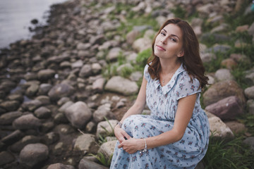 A beautiful young smiling brunette woman with long hair in a long blue dress sits by the lake or river on a warm summer day.