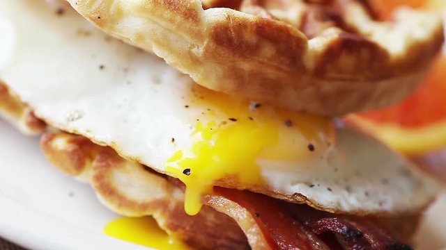 Peppered fried egg yolk drips slowly from a bacon and waffle sandwich