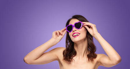 Naklejka premium valentines day, beauty and people concept - happy smiling young woman with pink lipstick and heart shaped sunglasses over ultra violet background