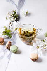 Fototapeta na wymiar Glass cup of hot green tea with french dessert macaroons, spring flowers white magnolia and cherry blooming branches over white marble texture background.