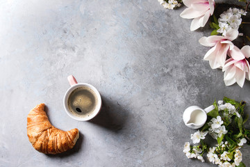 Pink mug of black espresso coffee, french croissant, cream and spring flowers magnolia, blooming cherry branches over grey texture background. Top view, space. Spring greeting card, wallpapers