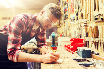 profession, carpentry, woodwork and people concept - carpenter with pencil writing to notebook at workshop