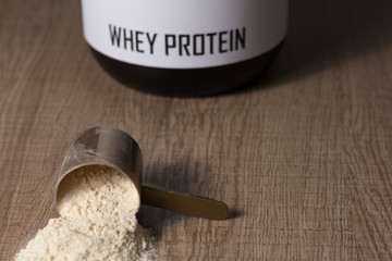 Whey Protein. Dropped scoop with vanilla powder flavour. Wooden table. Black jar behind