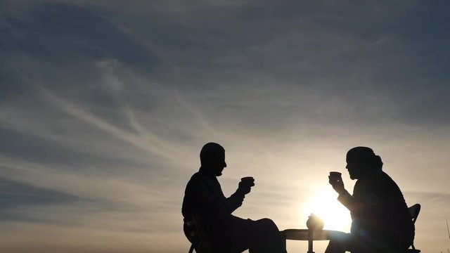 2 men silhouettes drink tea and communicate outdoors at sunset