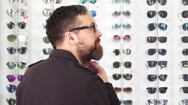 Rear view shot of a bearded mature man examining sunglasses for sale on the display at the opticians store smiling to the camera over his shoulder consumerism buying shopping retail.