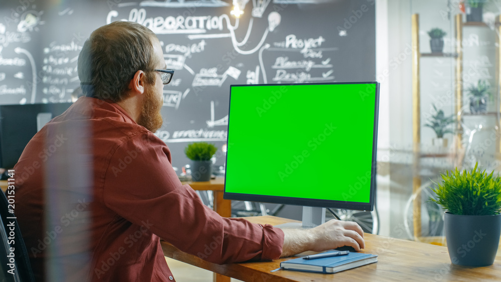 Poster Man Works on the Personal Computer with Green Mock-up Screen. In the background Creative Office with Chalkboard Wall.