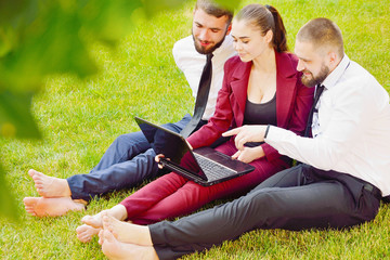 Young office workers with bare feet sit on a green lawn with a laptop and talk. Life style. Teamwork. Guys and the girl. 3.