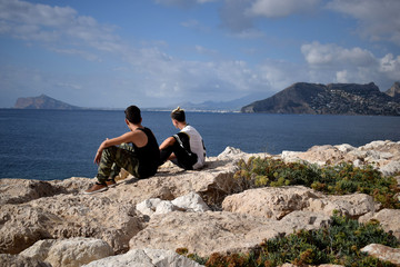 Fototapeta na wymiar Two young guys sit on the edge of the cliff and look at the beautiful view of the Mediterranean sea and mountains