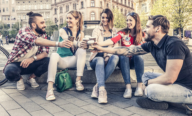 young cheerful people sitting in the street after work in the city center. they are toasting with...
