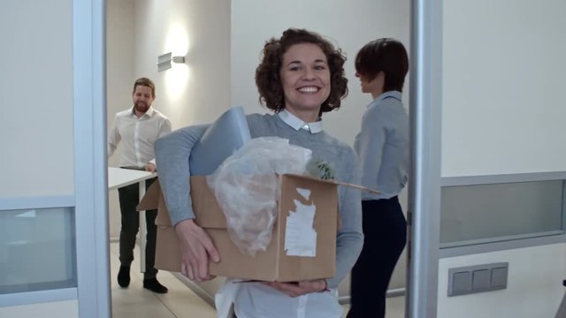 Young woman smiling, holding box with belongings and taking keys while walking into new office; male colleagues carrying table together