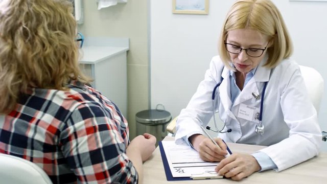Tilt up of cheerful female physician writing down symptoms of obese female patient