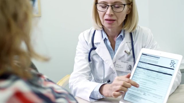 Tilt down of mature female doctor holding tablet and showing test results to unrecognizable patient: she pointing at data and explaining diagnosis; name Helen Brown in used in fictitious manner