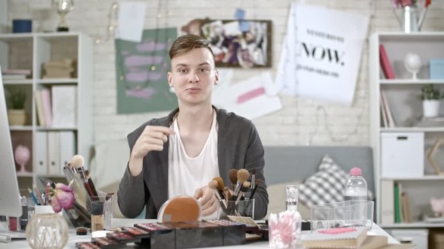 PAN of cheerful teenage boy looking at camera and showing brow brush to his viewers, then explaining how to use it and giving advice while filming makeup tutorial