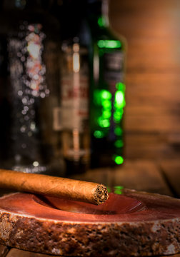 Cigar On Ashtray, With Liquor Alcohol Bottles In The Background Out Of Focus