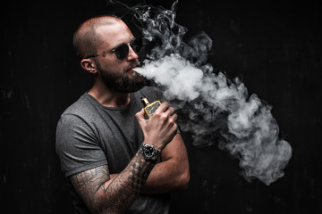 Men with beard in sunglasses vaping and releases a cloud of vapor on dark moody studio background.