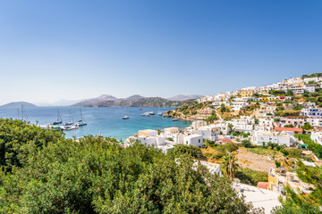 Fototapeta na wymiar Beautiful sunny greek village town harbor view to the aegean blue sea with crystal clear water boats cruising surrounded by hills mountains windmills on top, Panteli, Leros, Dodecanese Islands, Greece