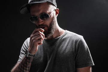 Pensive Black bearded male dressed in a grey shirt, sunglasses and baseball cap vaping. man in...