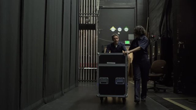 Two stage workers wheeling equipment trolley towards camera, backstage