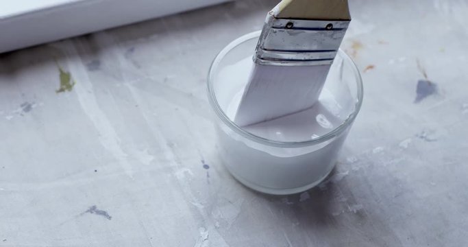 ECU detail artist dips broad brush into a pot of white gesso to use when priming canvas for oil painting. Hand-held 4K recorded in slow motion at 60fps.