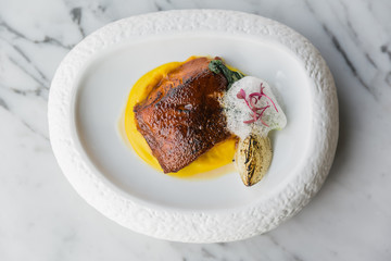 Top view of pan seared trout. served with yellow sauce, foam amd roasted sliced onion on curved white plate. 