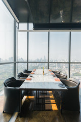 Six people dinner table with plates, knives, forks, wine glasses, glasses and napkins on marble table top with Bangkok cityscape in background.
