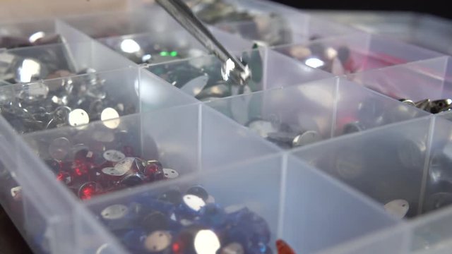 Multicolored decoration rhinestones in a plastic box. Woman is taking one with tweezers.